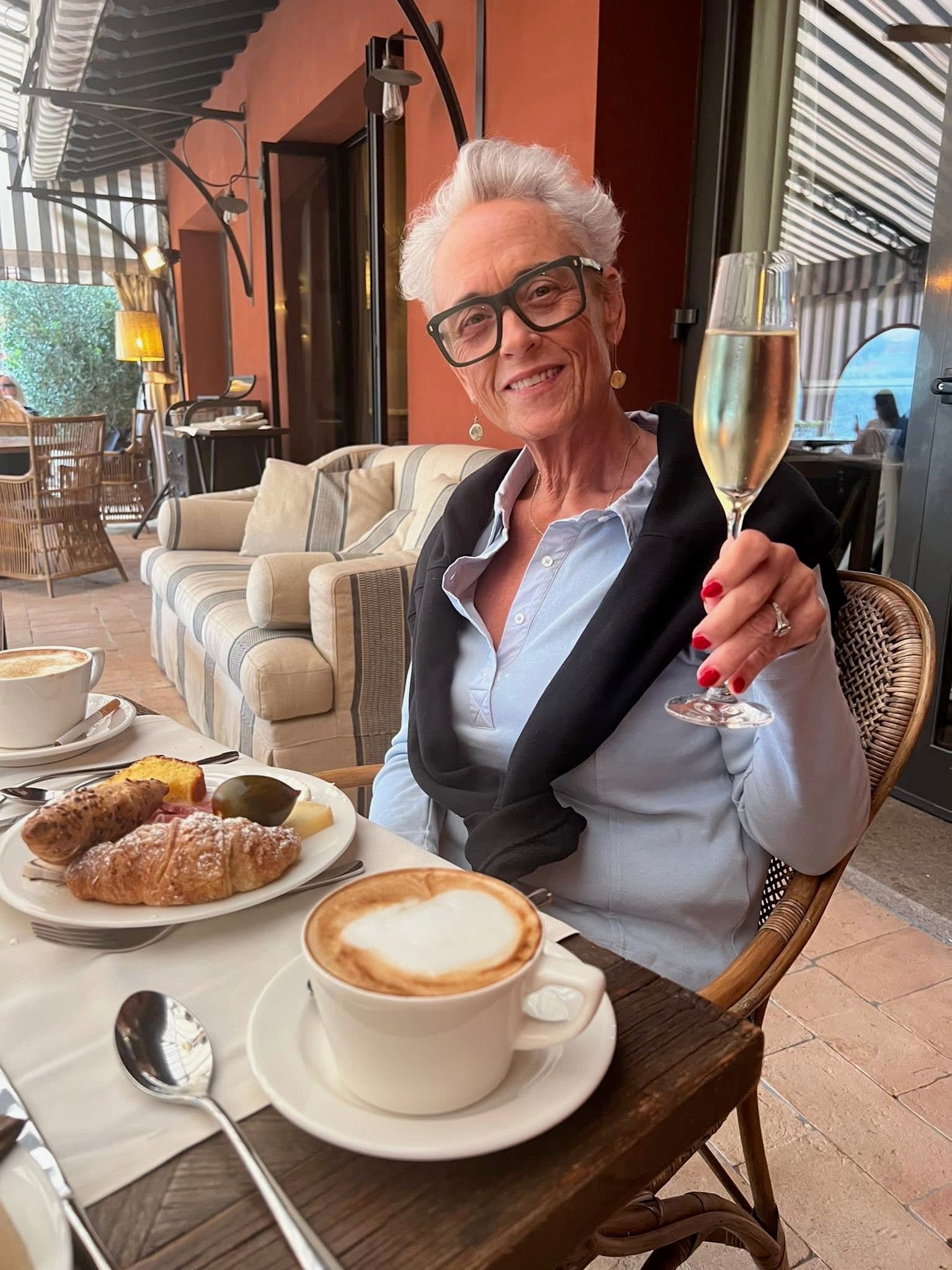 Smiling woman with white hair holding a champagne flute at an outdoor cafe table.