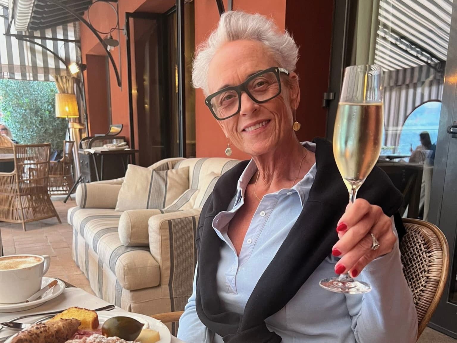 Smiling woman with short white hair and black glasses holding a champagne flute outdoors.