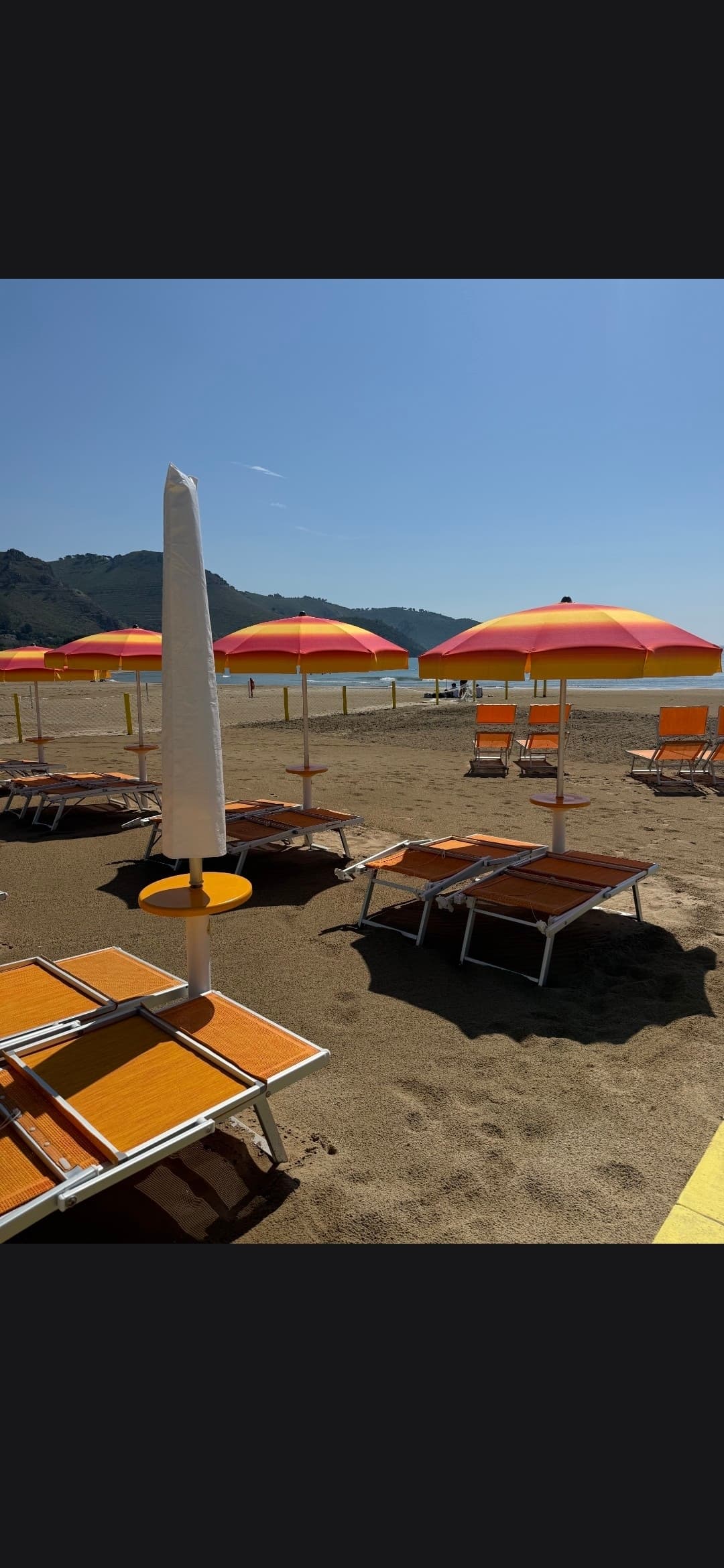 Rows of orange striped umbrellas and lounge chairs on a sandy beach with coastal mountains.