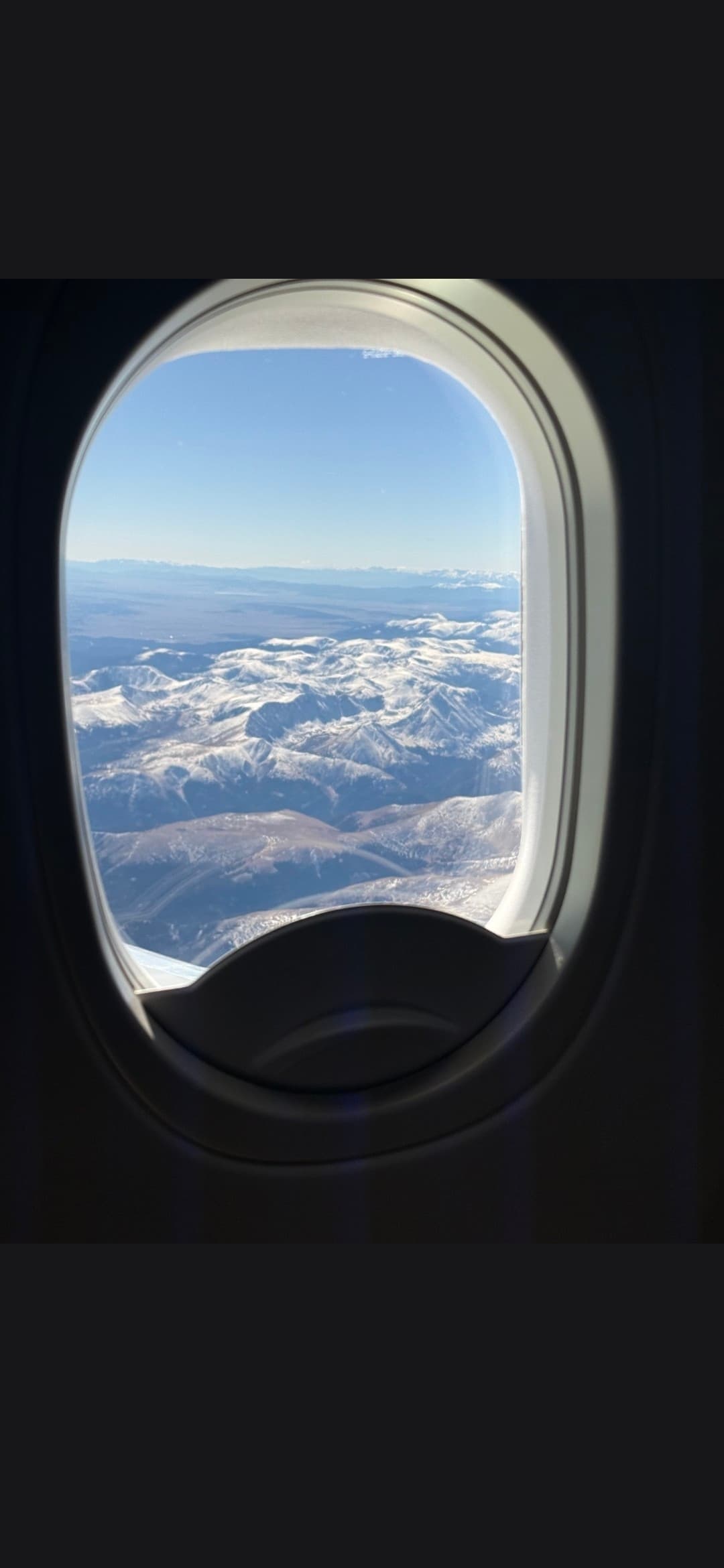 Aerial view of rugged, snow-capped mountains through an airplane window under a clear sky.