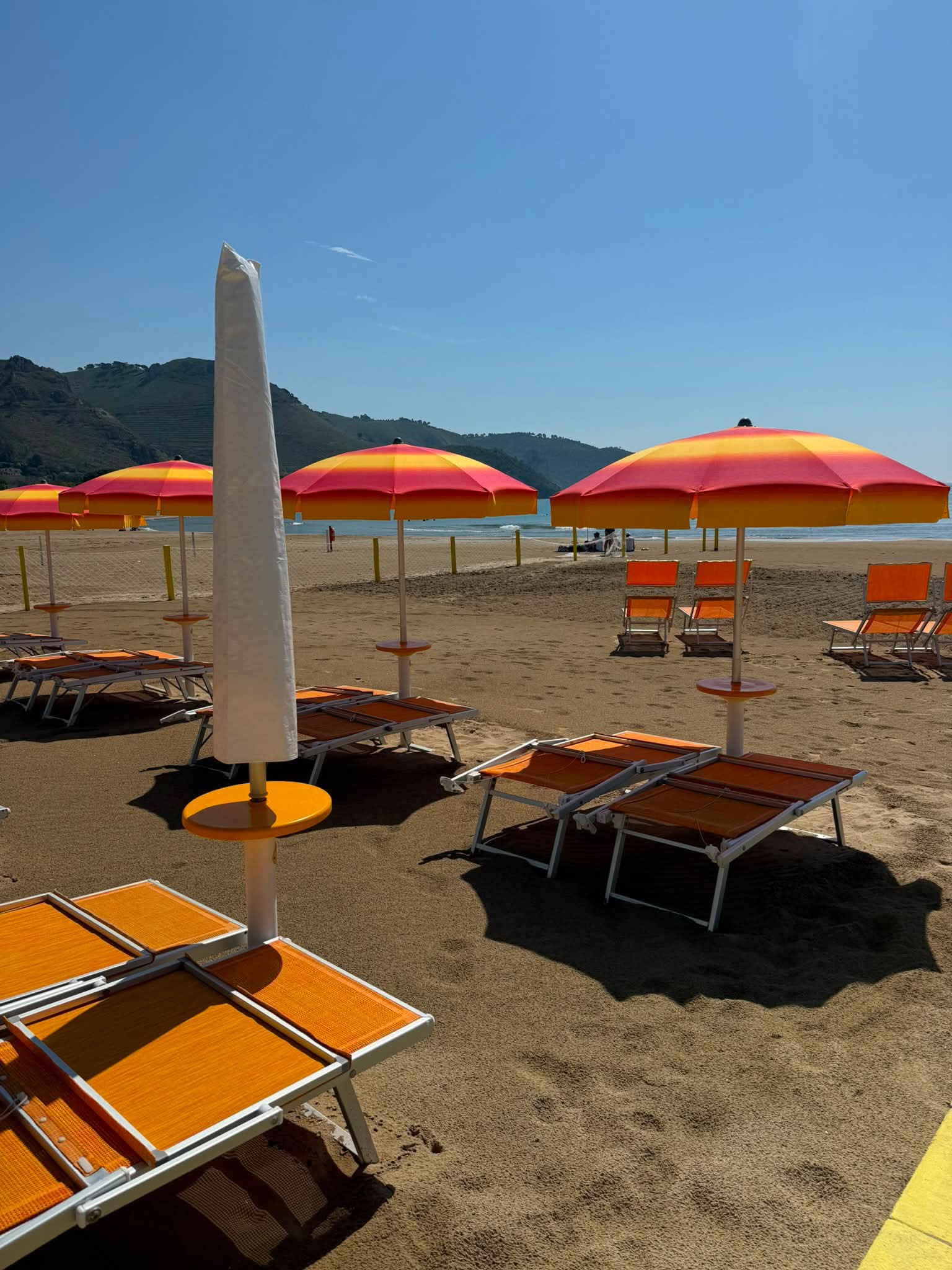 Orange and yellow striped beach umbrellas and sunbeds on a sandy shore with distant mountains.