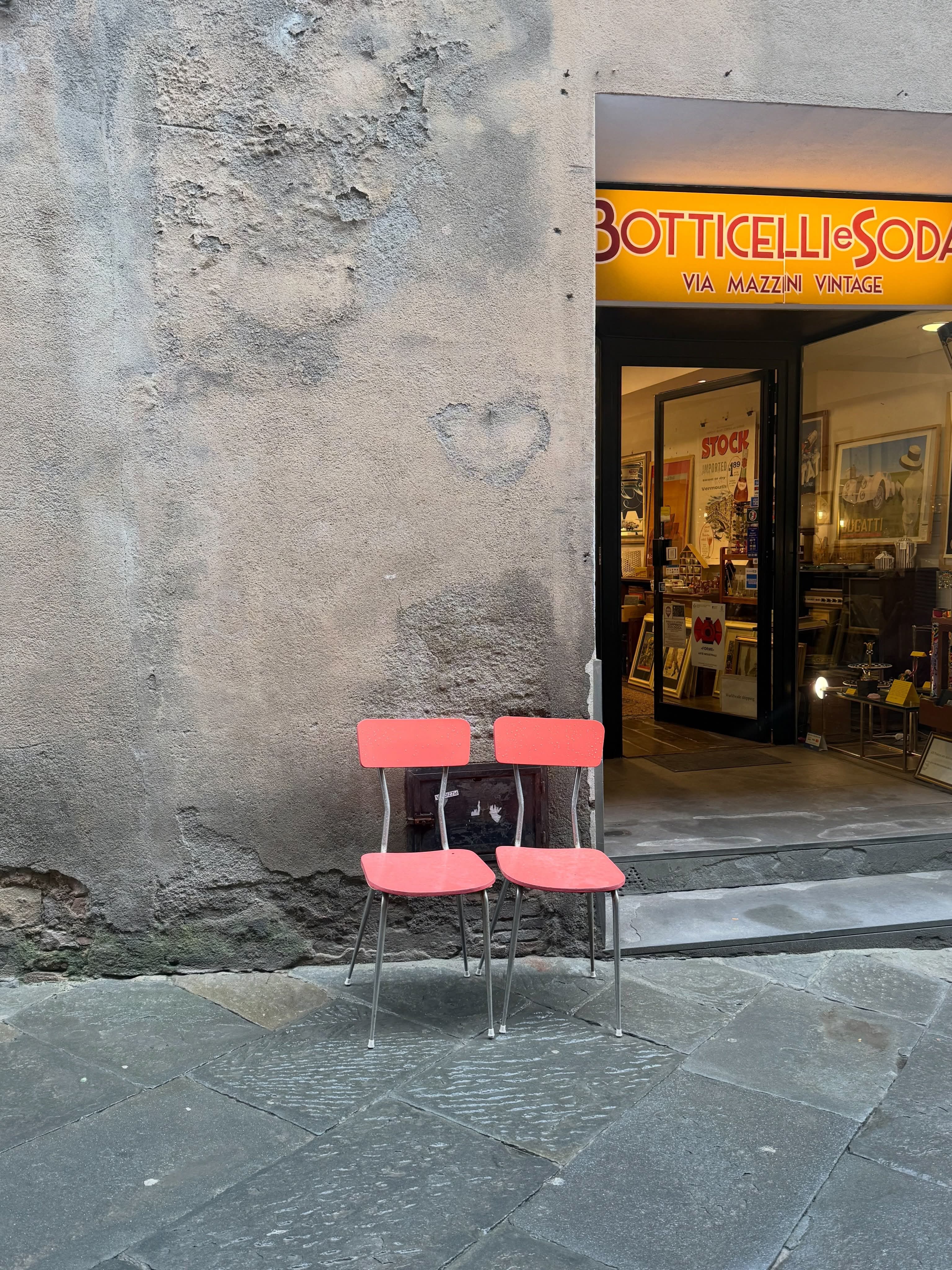 Two pink chairs against a weathered wall with a heart, beside a vintage shop.