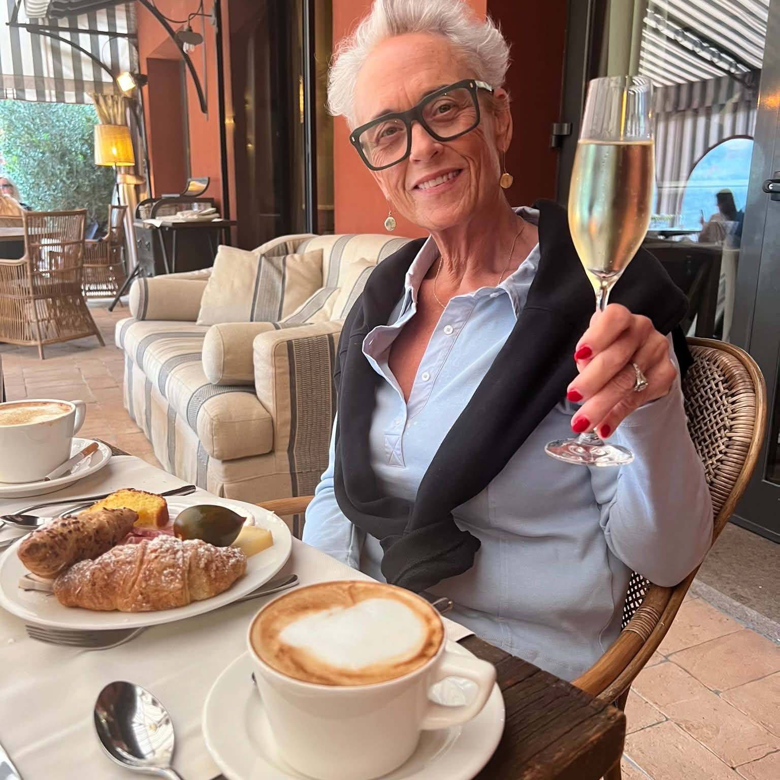 Smiling woman with short white hair raises a champagne flute over a breakfast spread.