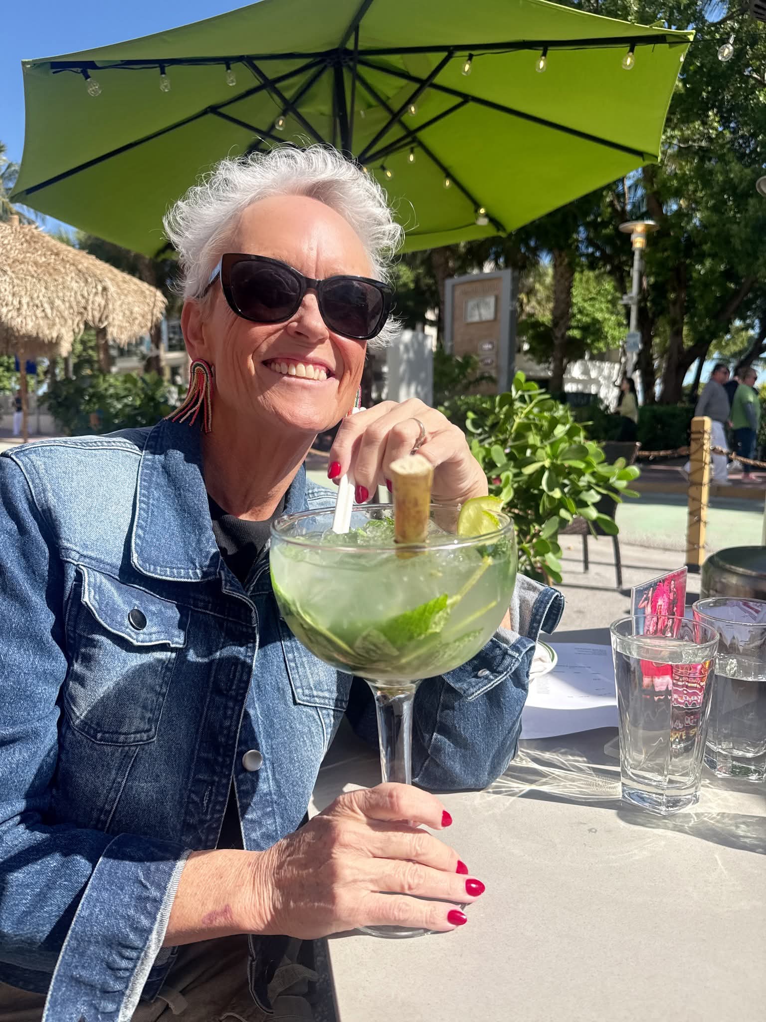 Smiling woman in a denim jacket holds a giant mojito under a sunny green umbrella.