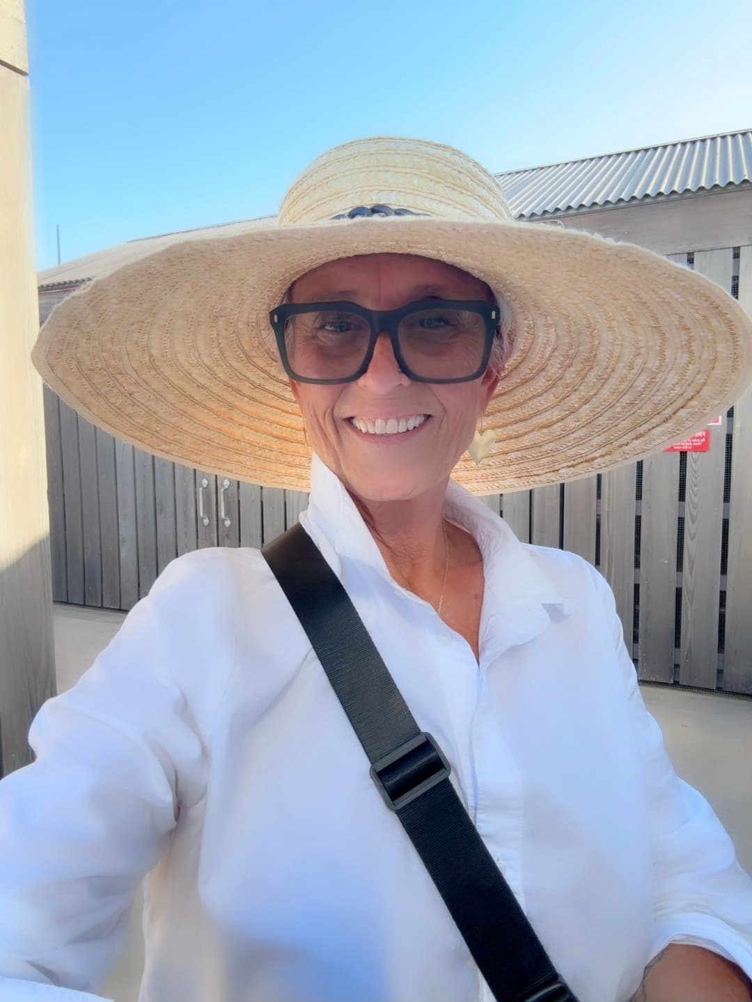 Smiling woman wearing a large wide-brimmed straw hat, black glasses, and a white shirt.
