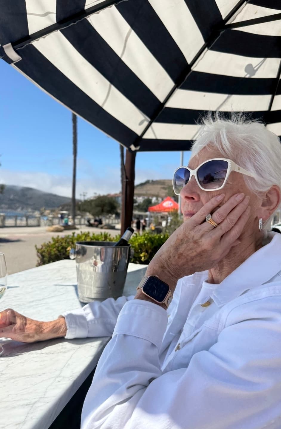 Elderly woman in white sunglasses sits at a seaside table under a striped umbrella.
