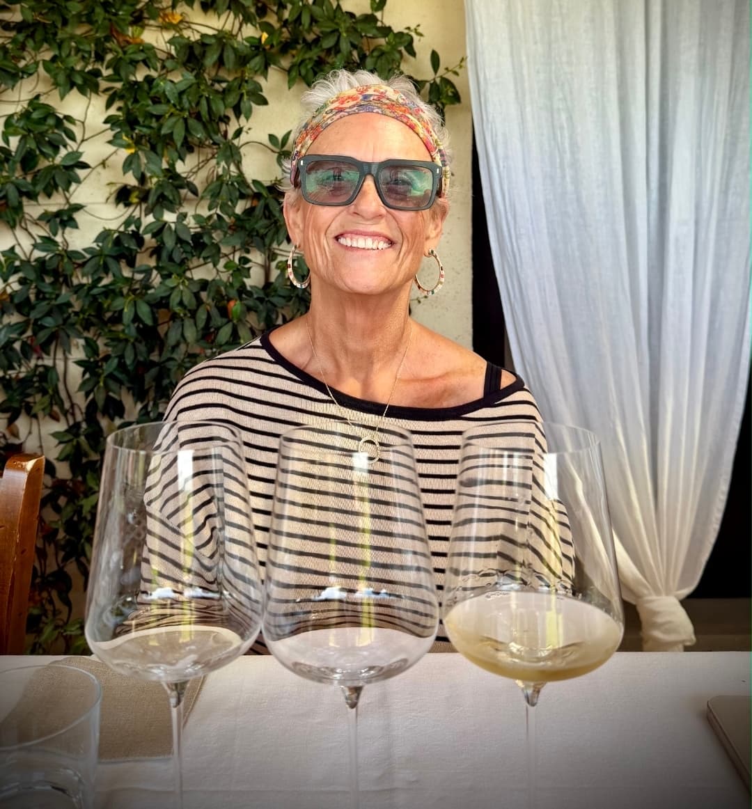 Smiling woman in a striped shirt and sunglasses sits behind three large wine glasses.